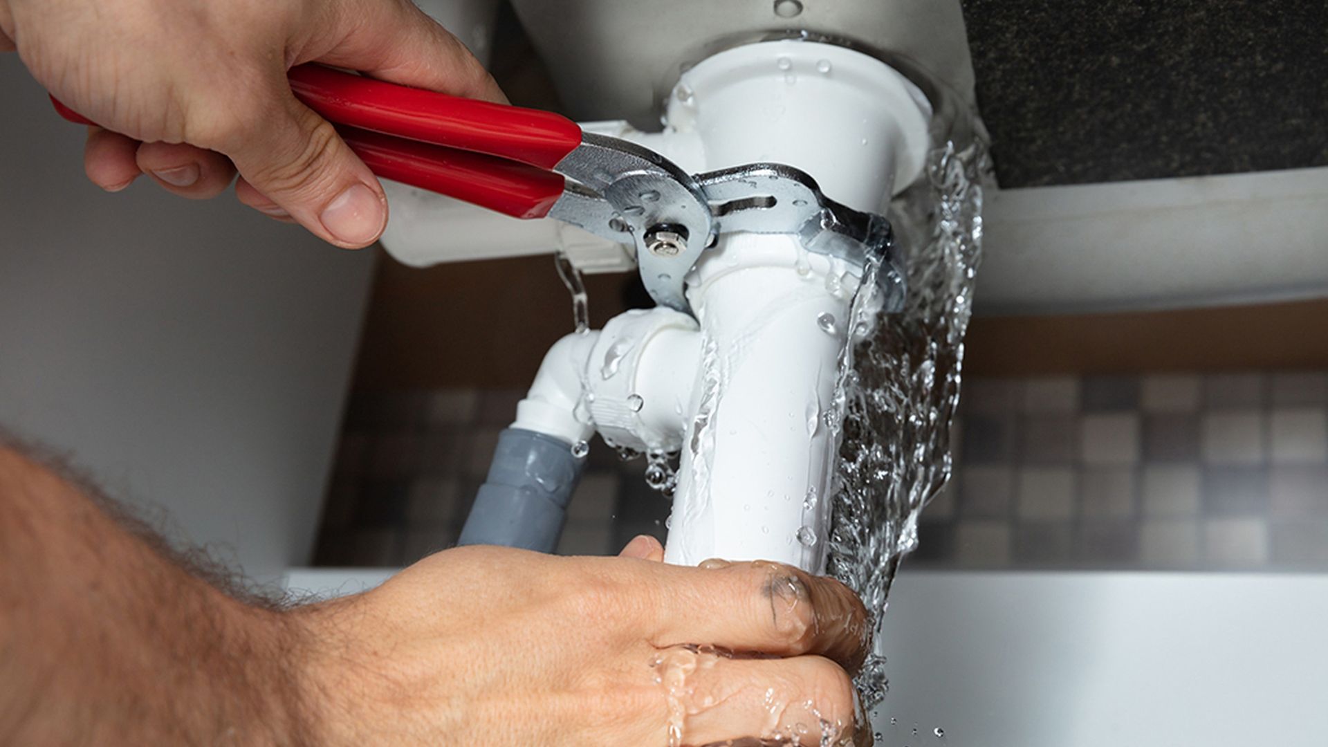 Plumber using wrench to repair leaking white pipe with water spraying