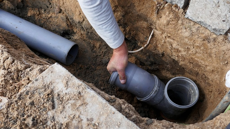 Worker installing underground drainage pipes in excavated dirt trench